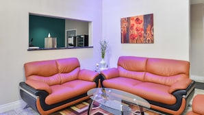 Un salon avec deux canapés en cuir orange, une table basse en verre, un vase avec des branches, un tableau, et un coin cuisine à l’arrière-plan.