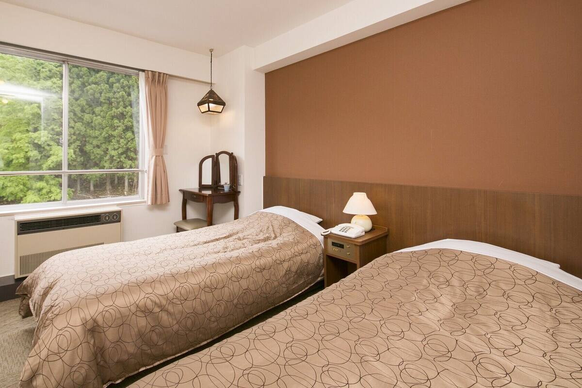 Habitación de hotel con dos camas, un escritorio, una silla, una lámpara y una ventana con vista a árboles.