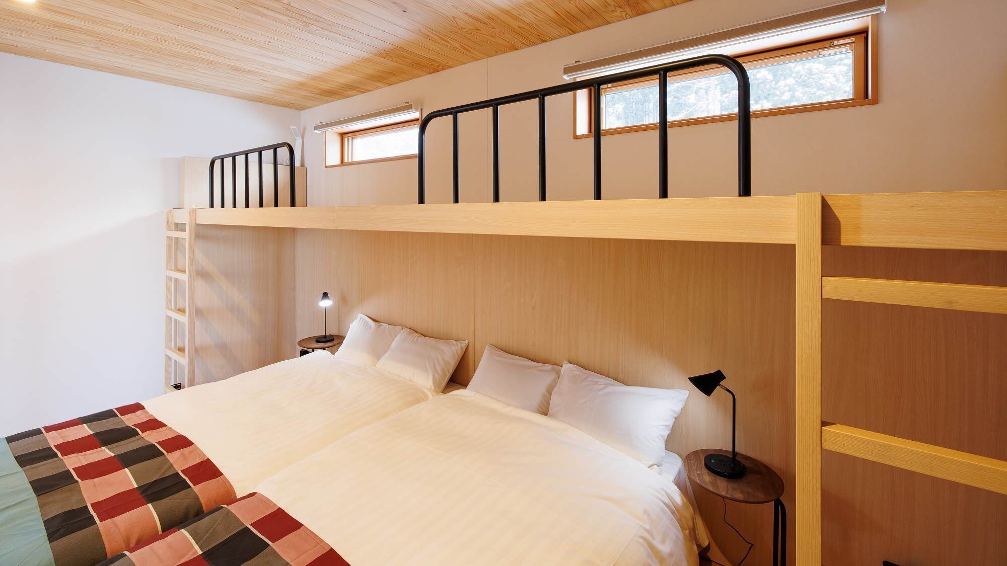 Uma cama beliche com cabeceira de madeira, uma mesa de cabeceira com abajur e uma janela com vista para árvores.