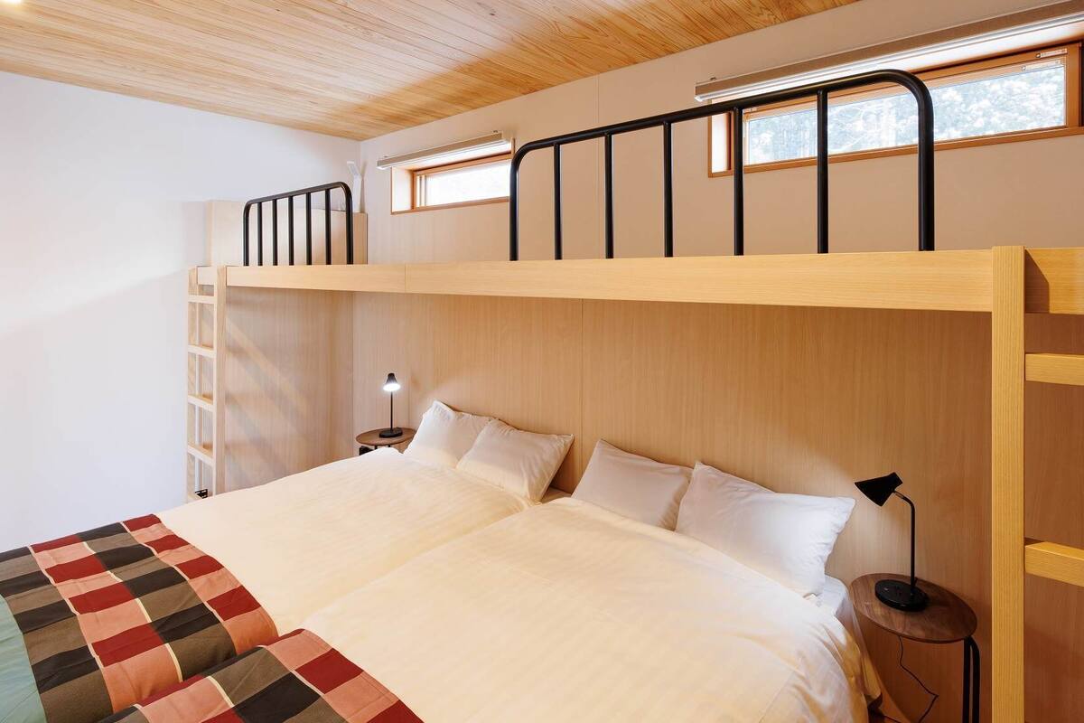 Una cama individual con cabecera de madera, una mesita de noche con lámpara, y una ventana con vista a árboles.