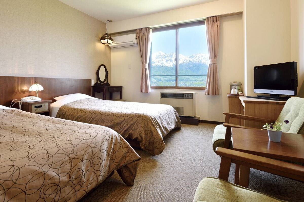 Habitación de hotel con dos camas, un escritorio con silla, televisor, ventana y vista a la montaña, además de un calentador.