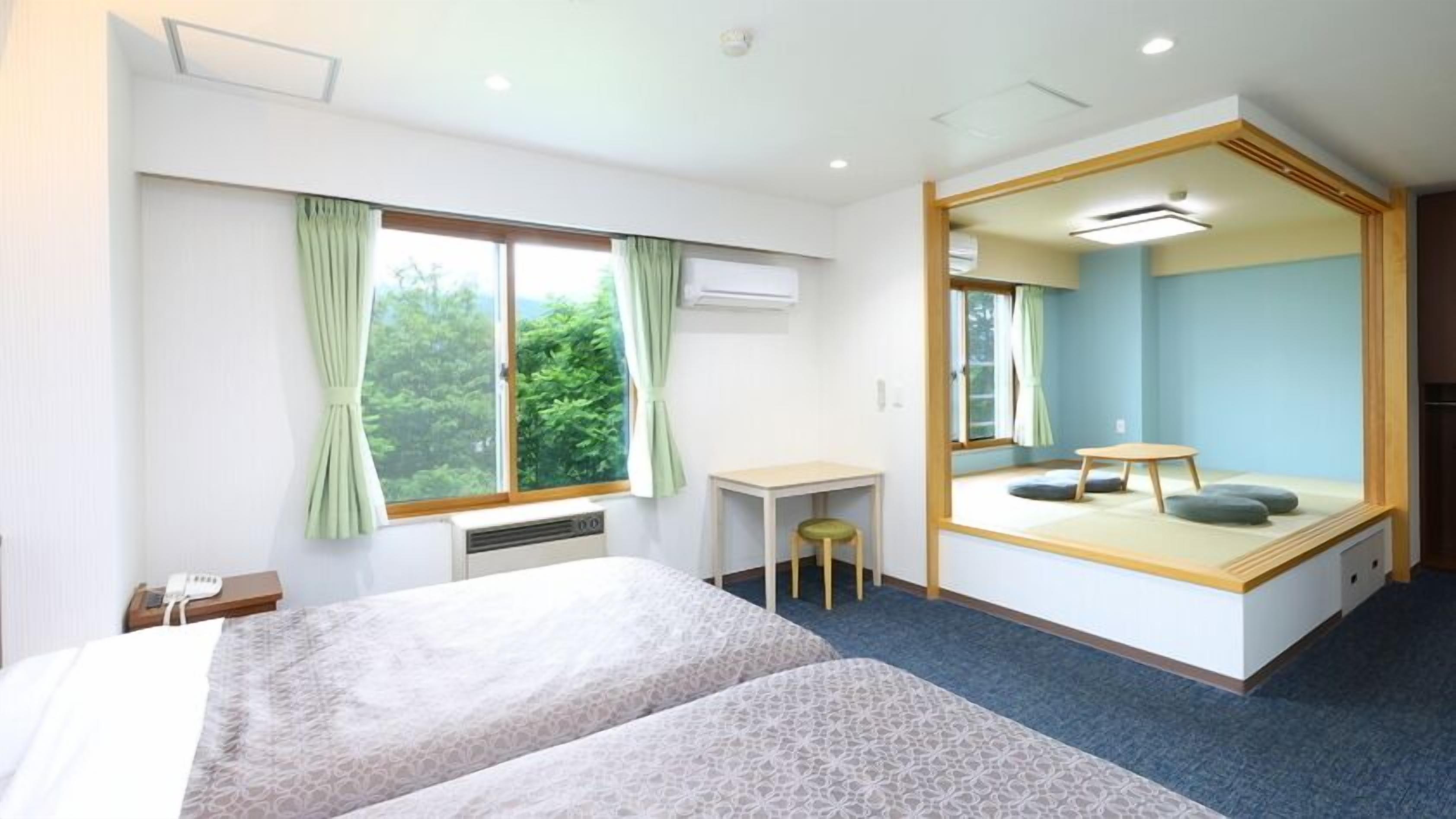 Quarto de hotel com uma cama, escrivaninha, cadeira, banquinho pequeno, espelho e vista para o verde lá fora.