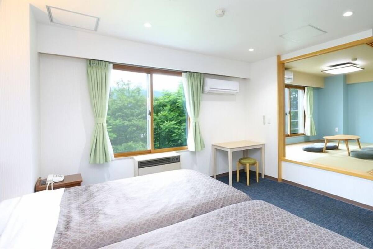 Habitación de hotel con cama, escritorio, silla, taburete pequeño, espejo y vistas a zonas verdes.