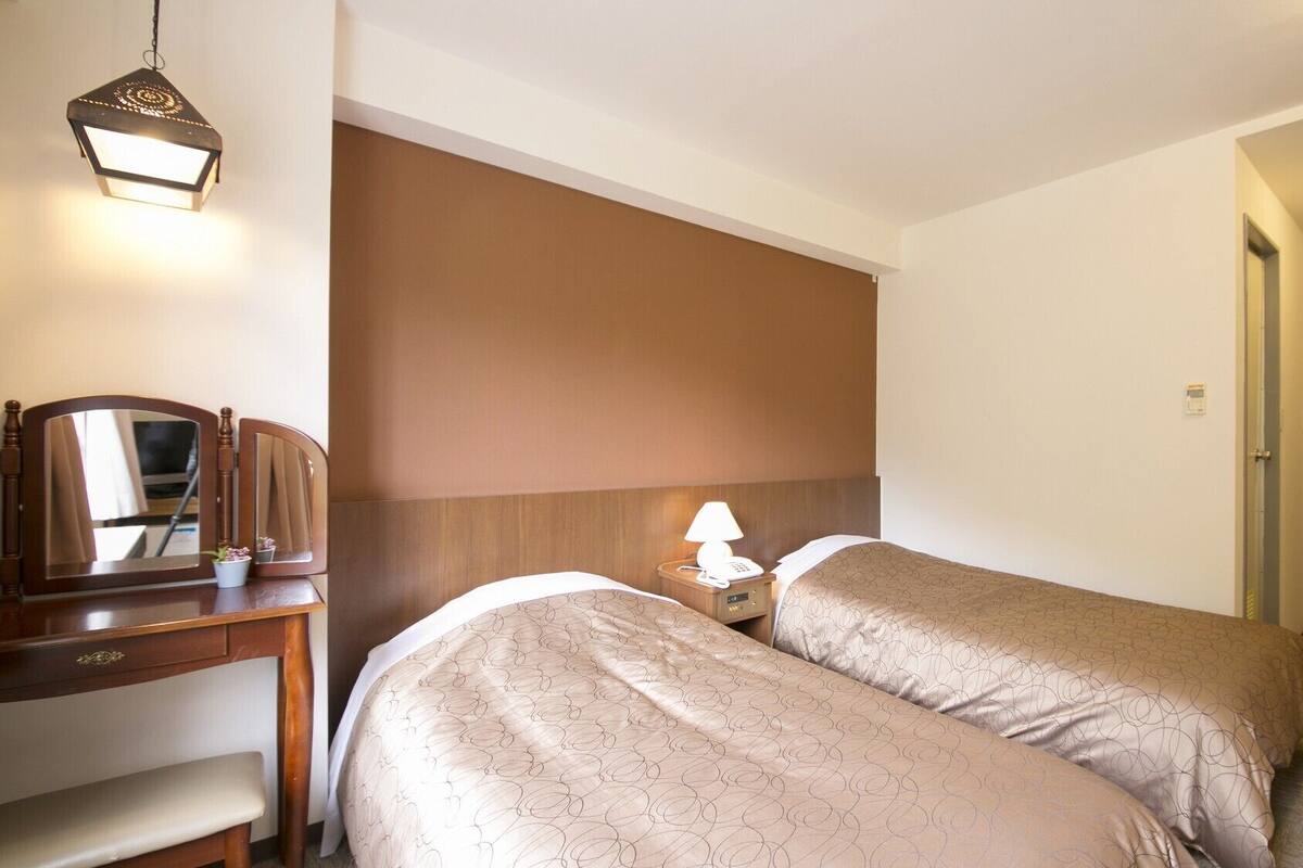 Habitación de hotel con dos camas, un escritorio con espejo, una mesita de noche con lámpara y una puerta de madera.