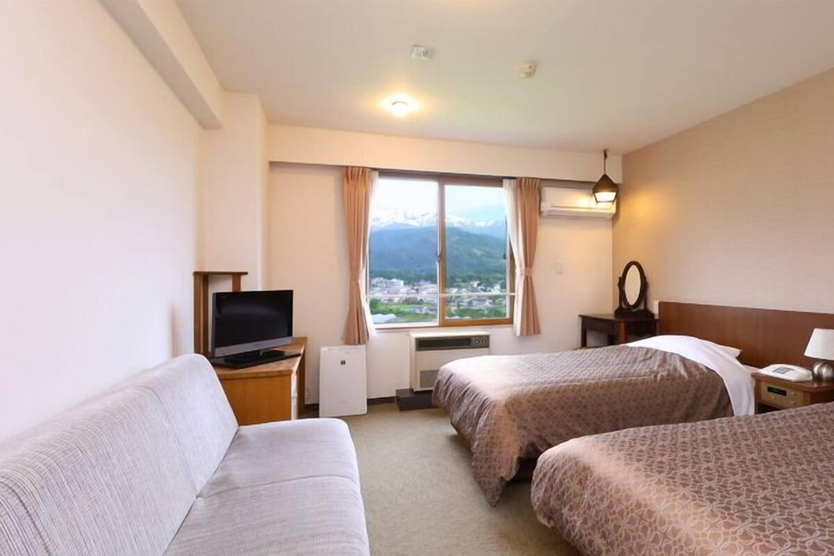 Habitación de hotel con dos camas, televisión, calefacción, ventana con vistas y una lámpara.