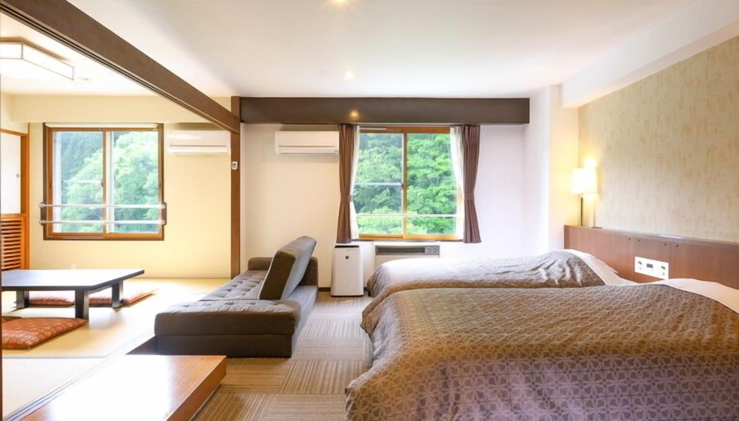 Quarto de hotel com duas camas, um sofá, uma mesa e vista para o verde pelas janelas.