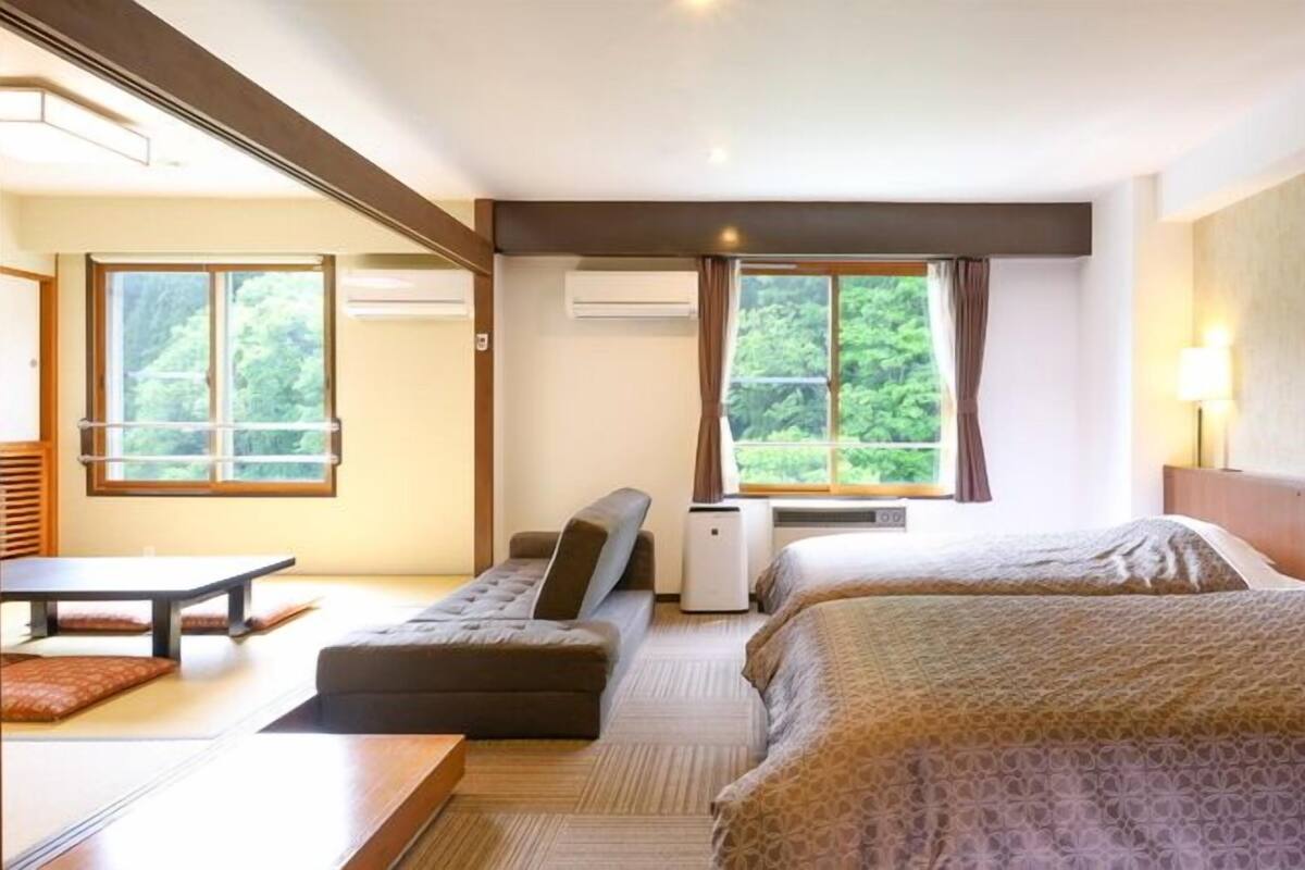 Habitación de hotel con dos camas, un sofá, una mesa y vistas a zonas verdes por las ventanas.