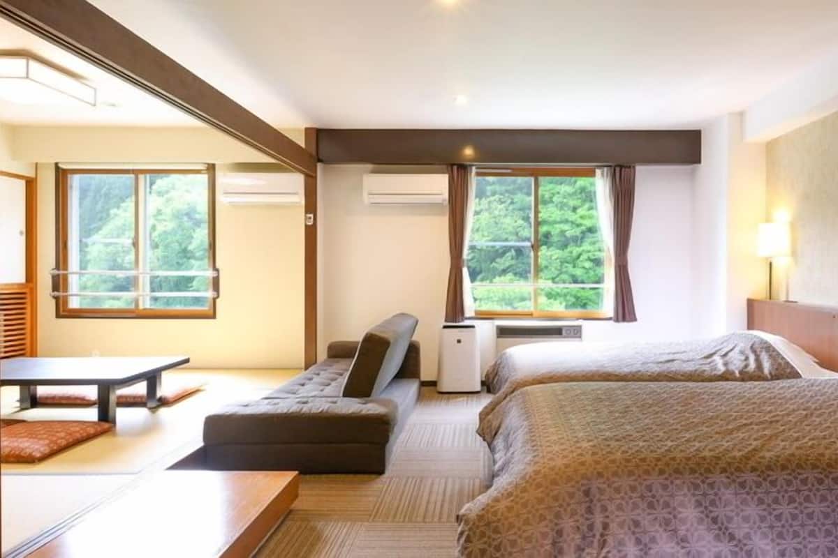 Habitación de hotel con dos camas, un sofá, una mesa y vistas a zonas verdes por las ventanas.
