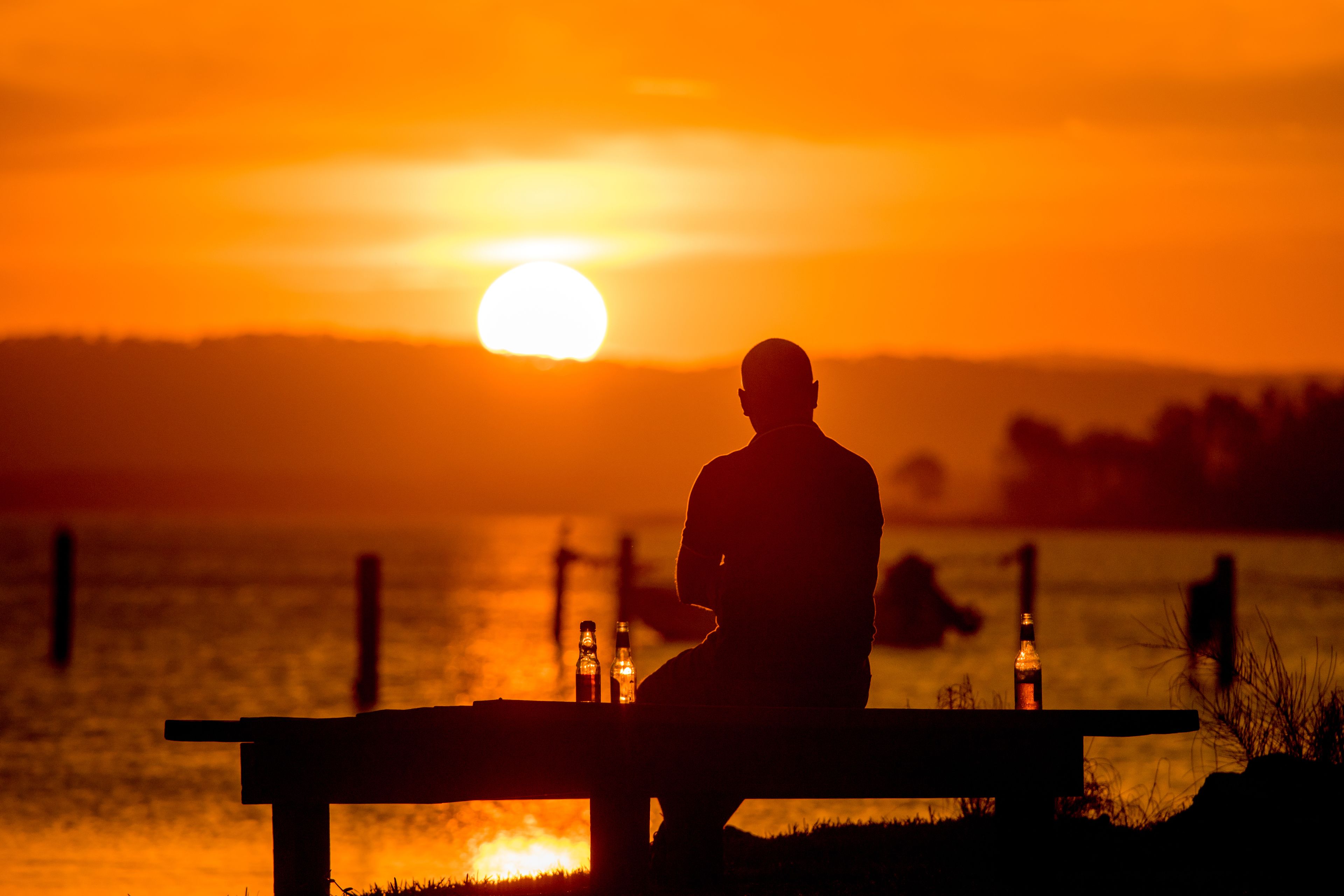A person sitting on a bench at sunset with bottles on it.