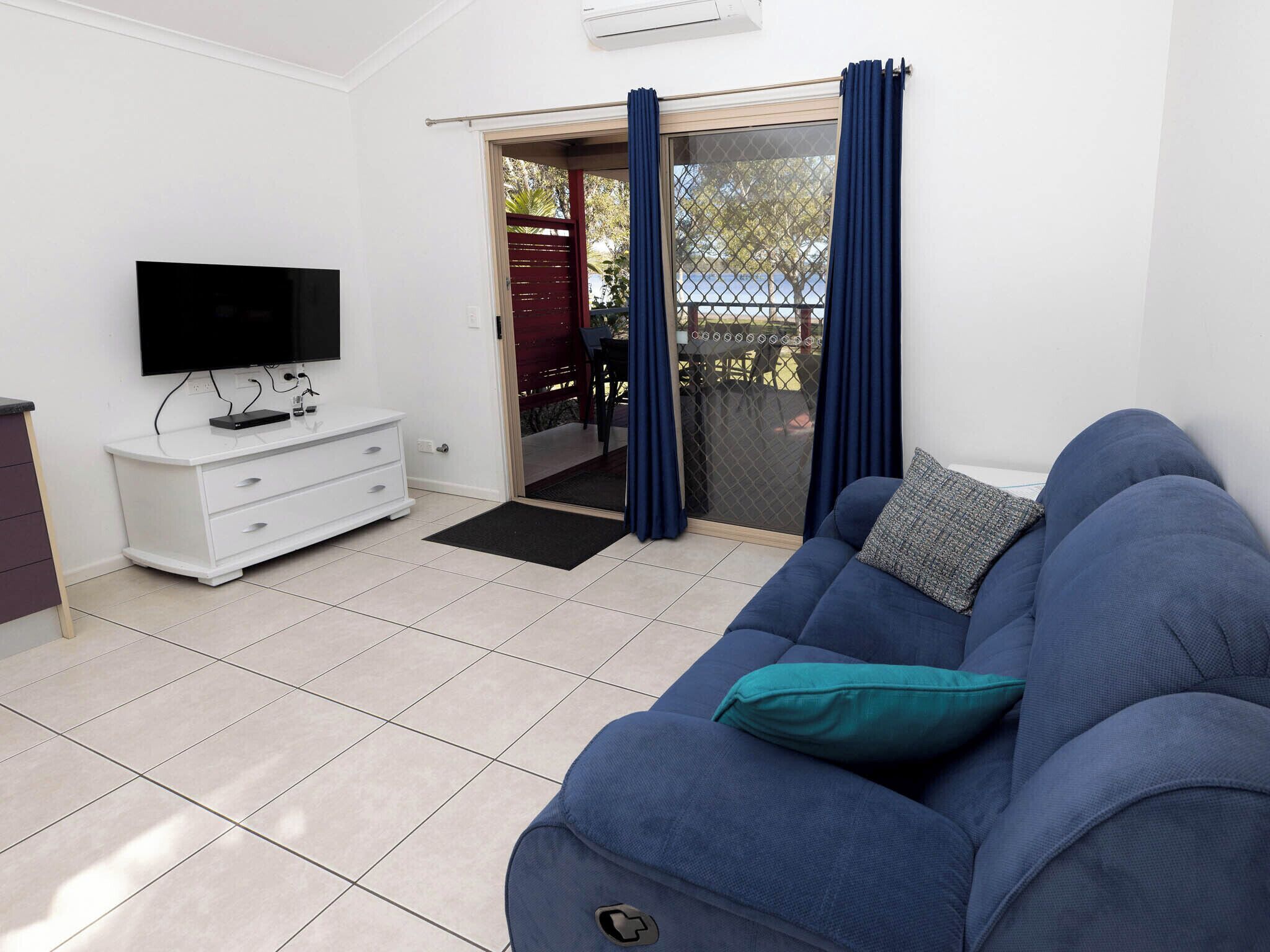 A modern living room with a white TV stand, a blue sofa, and a sliding glass door leading to an outdoor area.