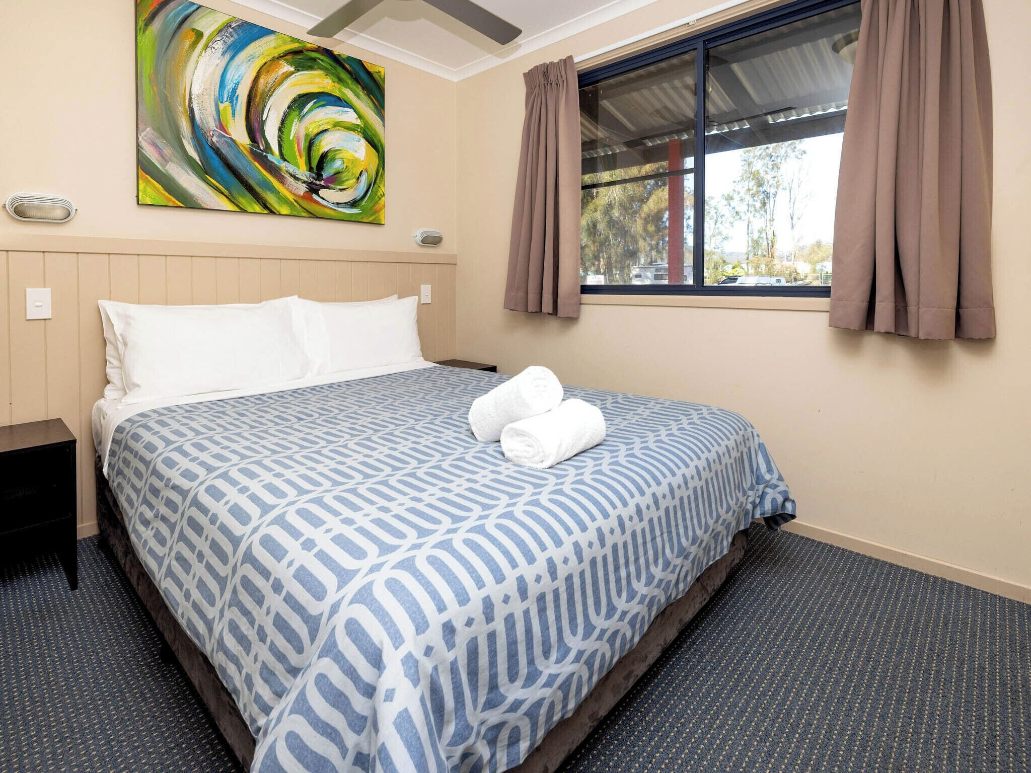 A hotel room with a bed, a painting on the wall, and a window with curtains.
