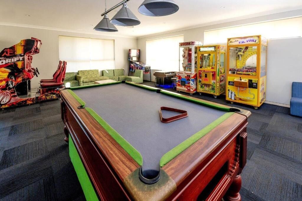 A pool table with green and red cushions, a sofa, and arcade machines in a room with carpeted flooring and a ceiling light fixture.