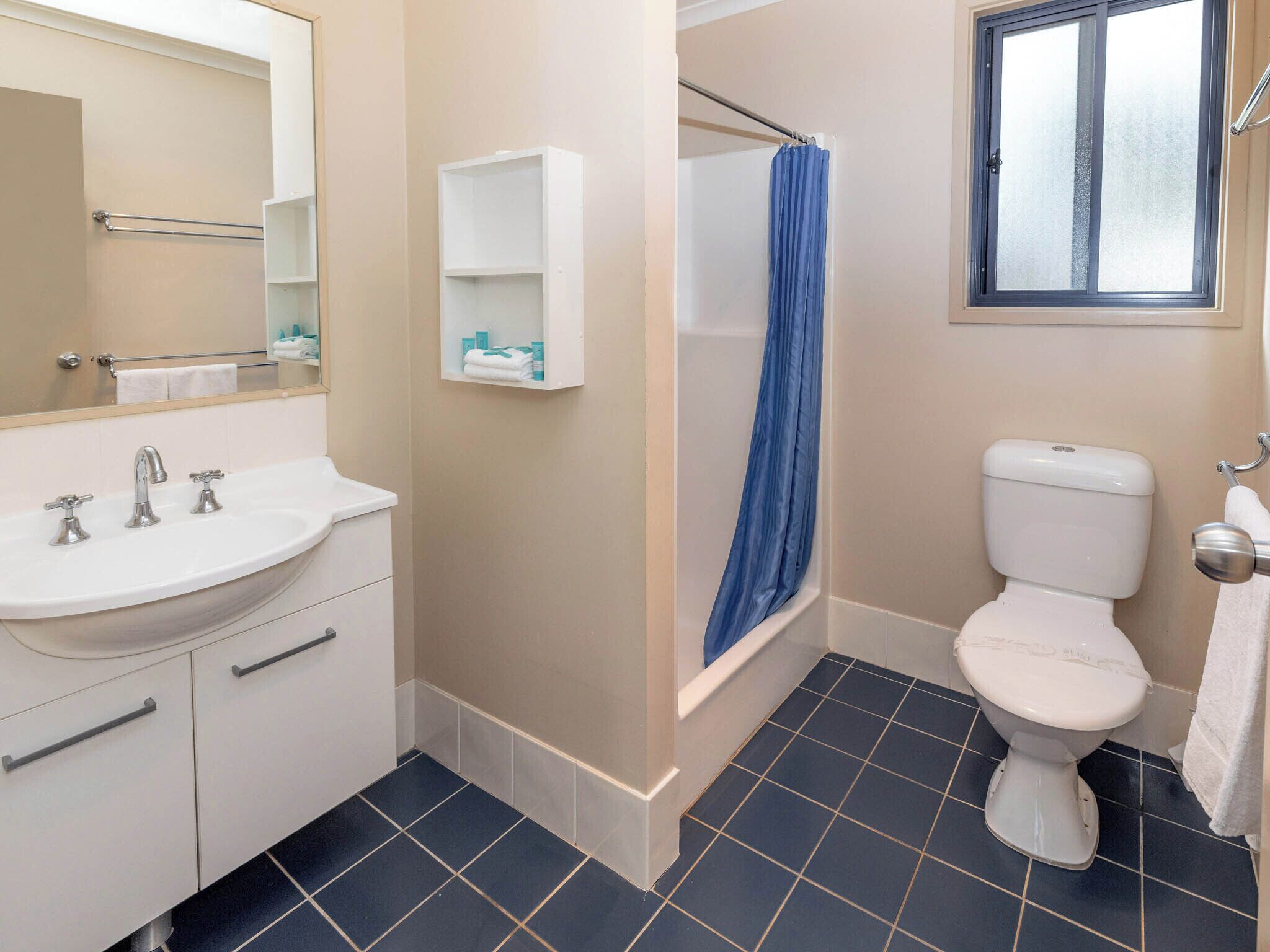 A bathroom with a white sink, two faucets, a mirror, a white toilet, a blue shower curtain, and a window with frosted glass.