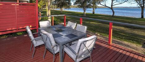 An outdoor deck with a table and chairs, overlooking a lake with trees and a red fence.