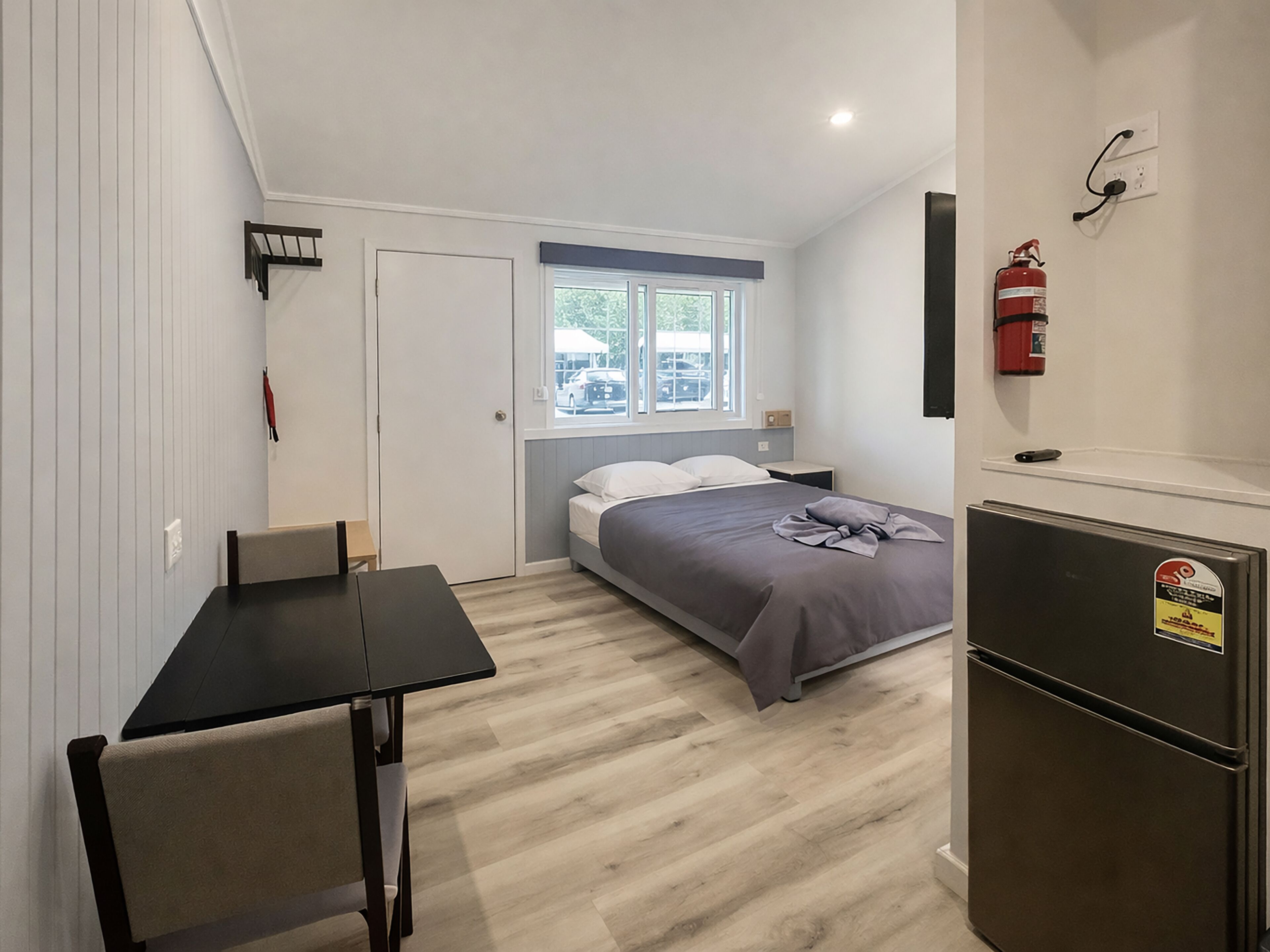 A compact bedroom with a bed, a small table, a refrigerator, and a window.