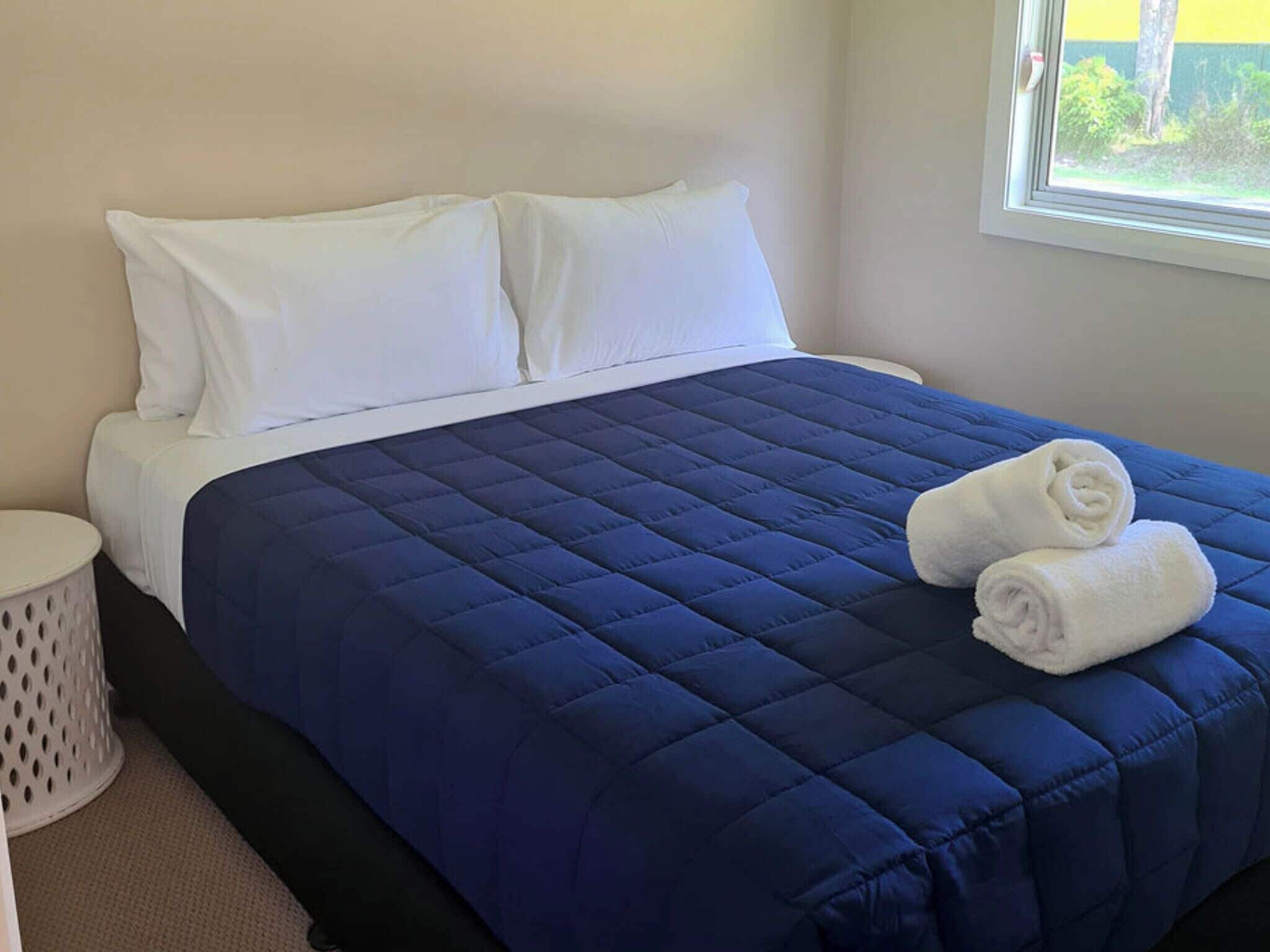 Een keurig opgemaakt bed met een blauwe deken, witte kussens en twee opgerolde witte handdoeken op het bed.