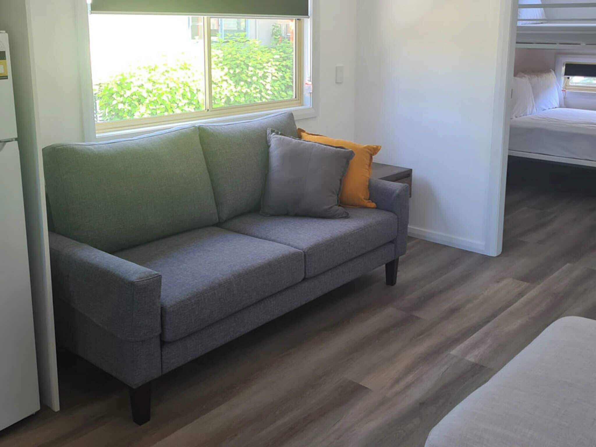 Un sofá gris con cojines en una habitación con suelo de madera y una ventana con vistas a la vegetación.