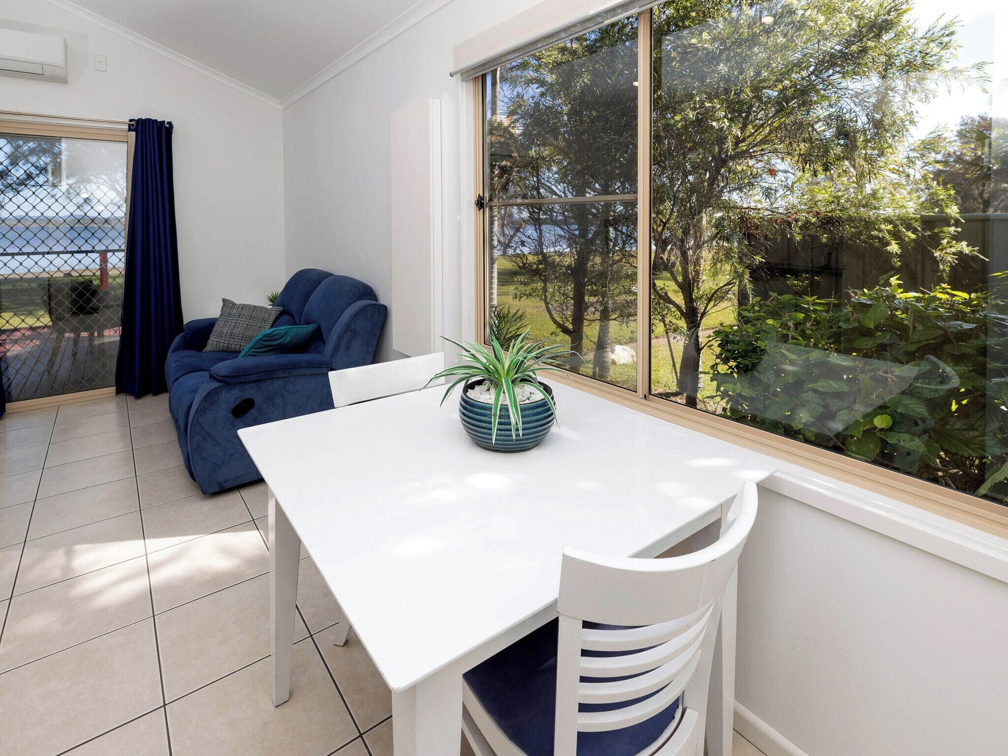 Un salon moderne avec une table blanche et un canapé bleu, une grande fenêtre donnant sur des arbres et une plante en pot posée sur la table.