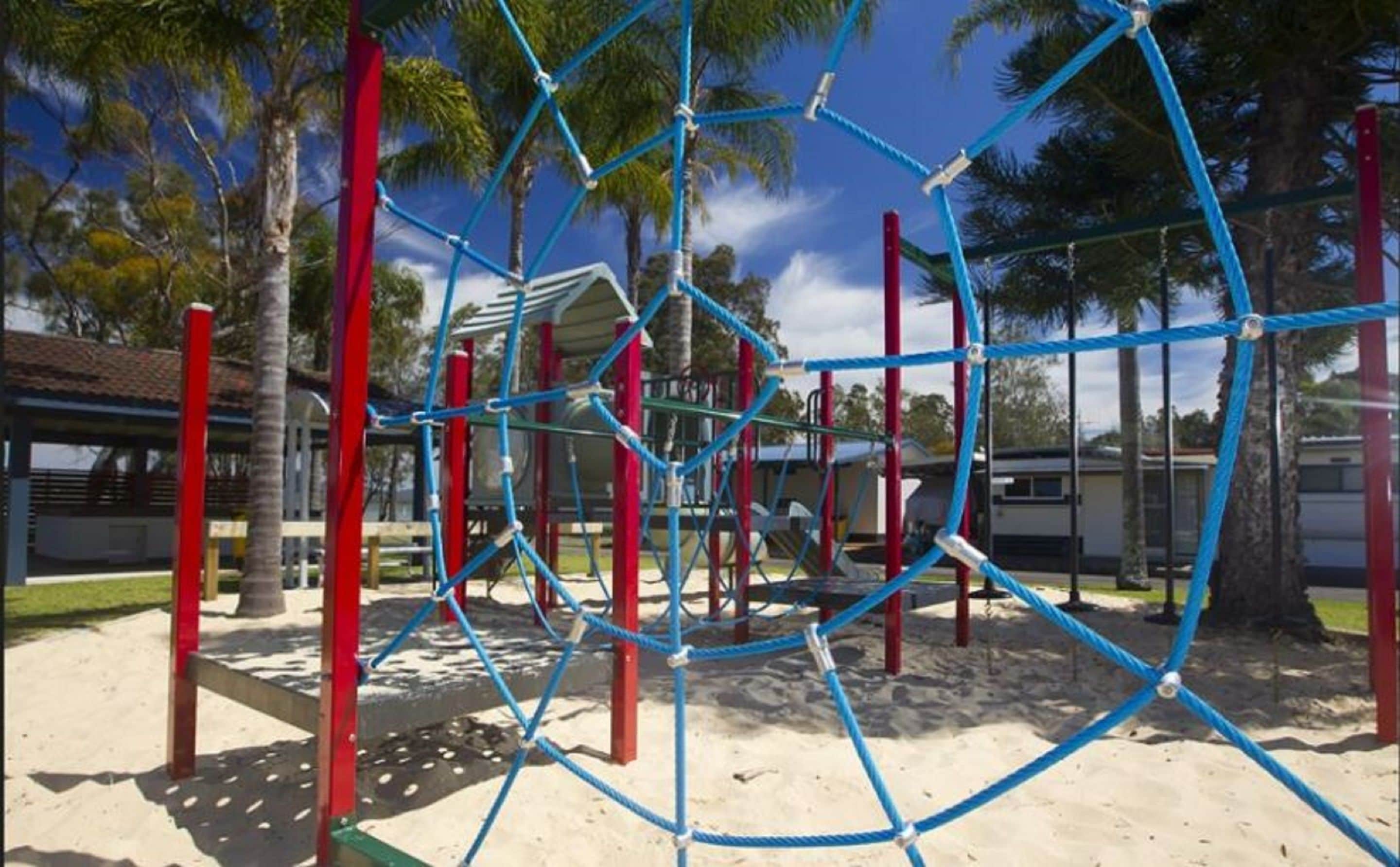 A playground with a climbing structure, red and blue poles, and a sandy ground.