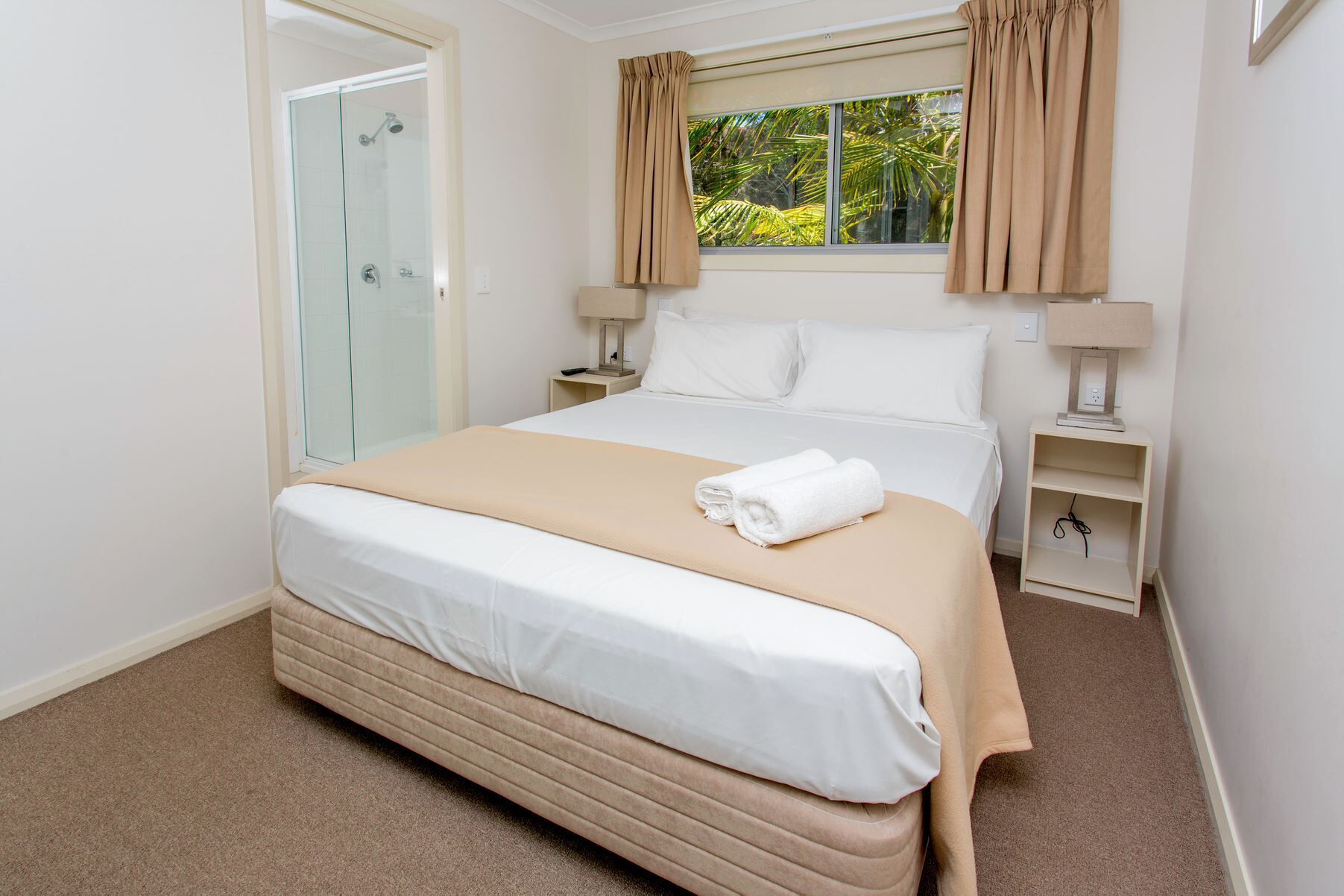 A hotel room with a bed, a nightstand, a lamp, a window with curtains, and a glass shower enclosure.