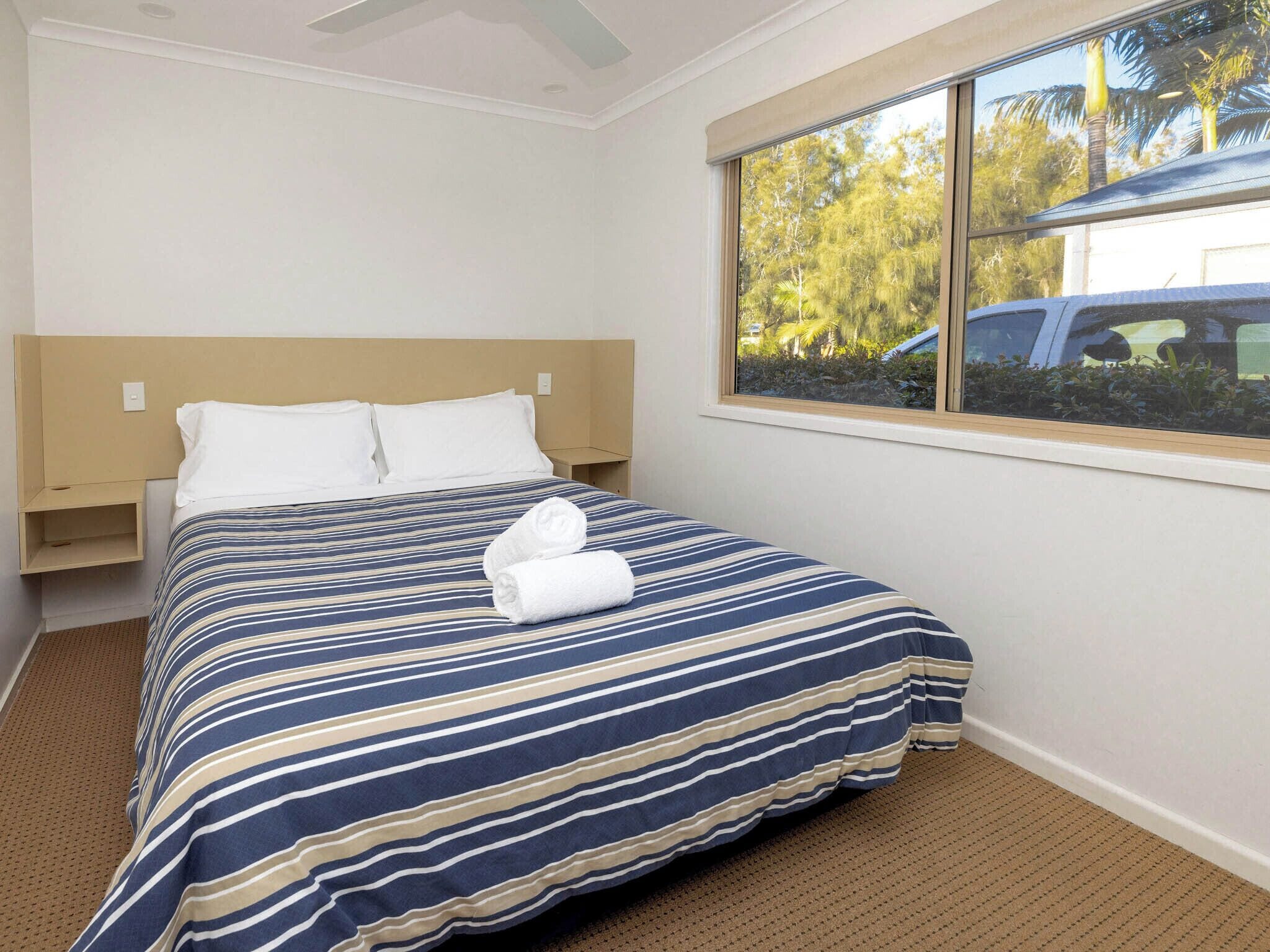 A hotel room with a large bed, a window with a view of trees and a parked car, and a ceiling fan.