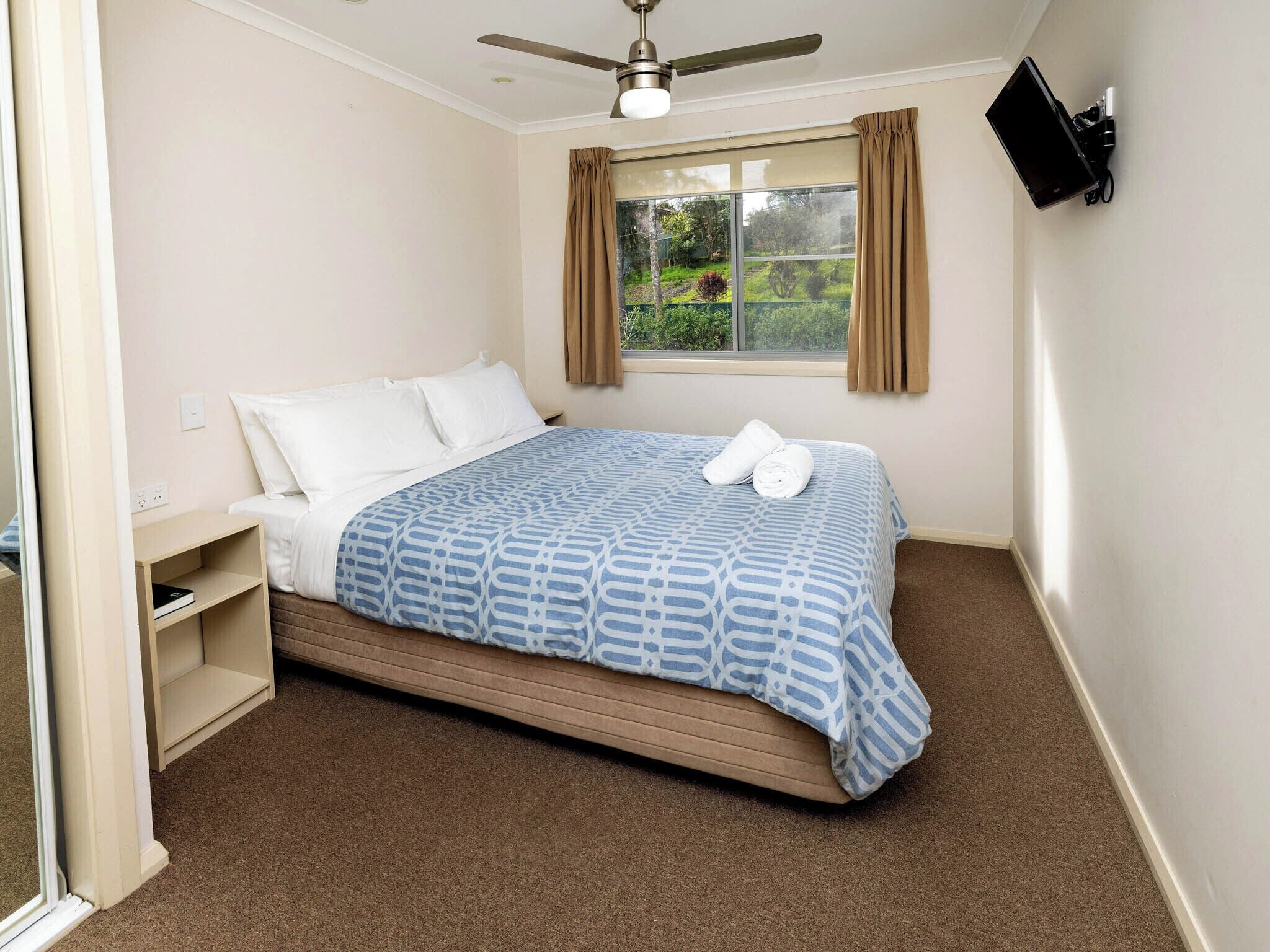 A bedroom with a bed, a ceiling fan, a TV mounted on the wall, and a window with curtains.