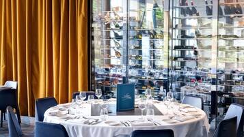 A restaurant with a round table set for dining, a menu stand, and a view of wine bottles through a glass wall.