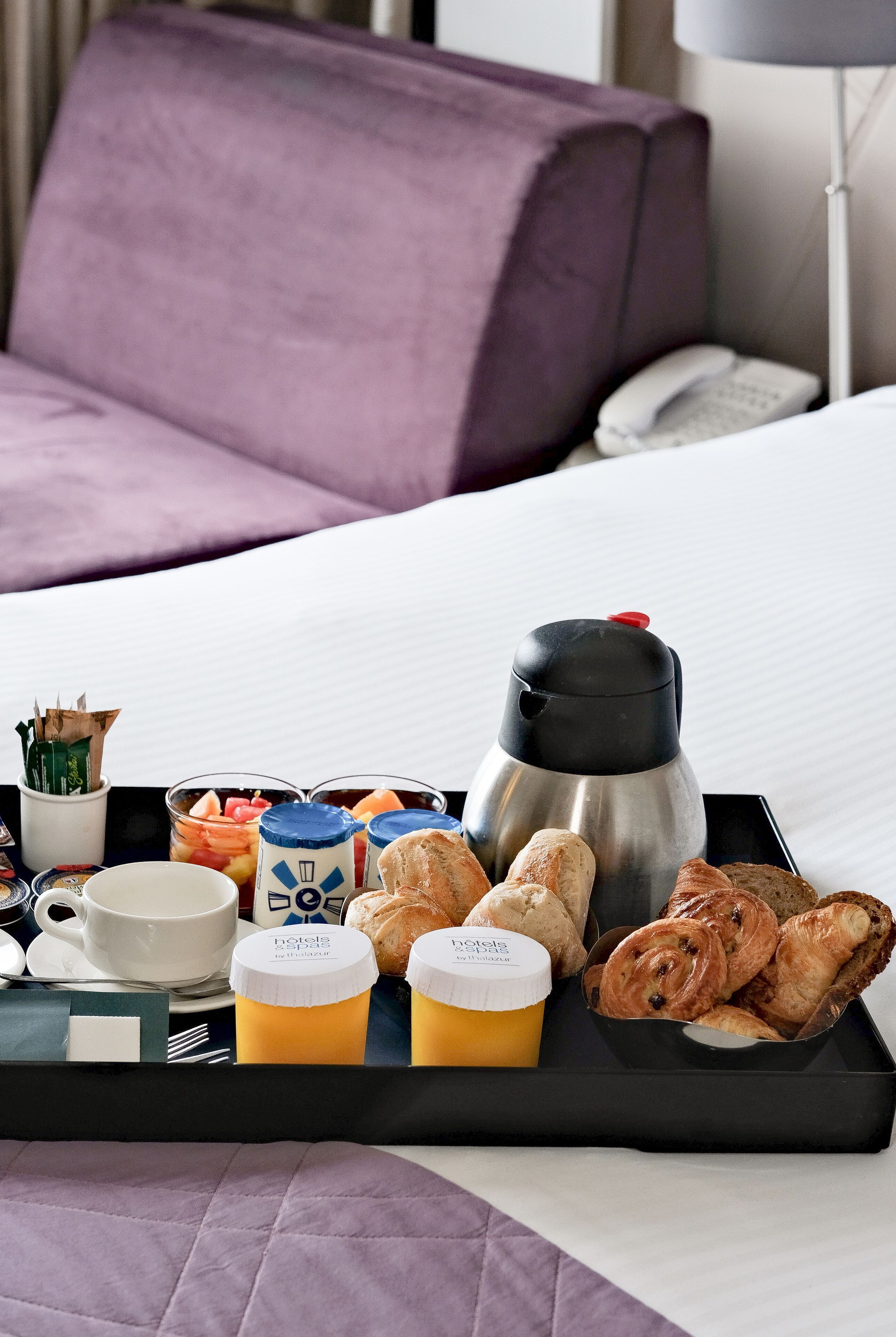 Un plateau de petit-déjeuner avec des viennoiseries, du café et du jus, posé sur un lit dans une chambre d’hôtel.