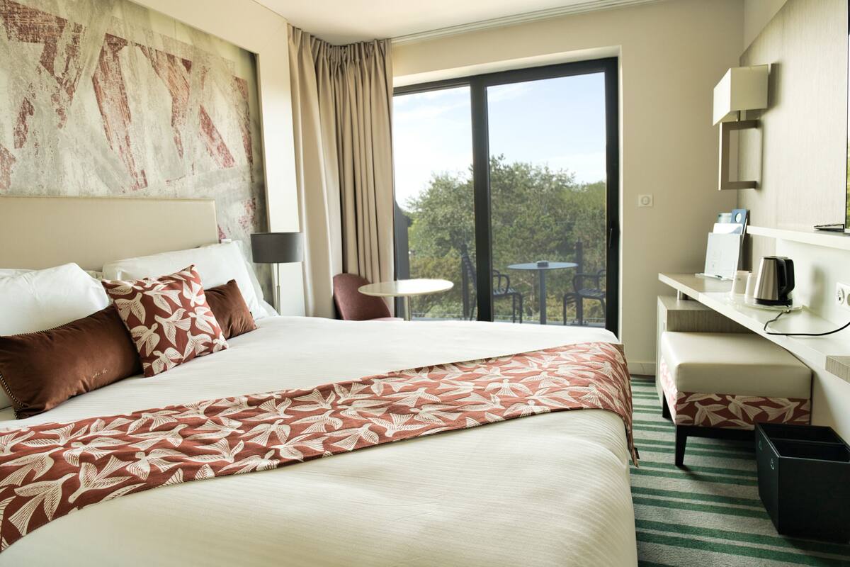 A hotel room with a large bed, a desk with a chair, a window with a view of trees, and a balcony.