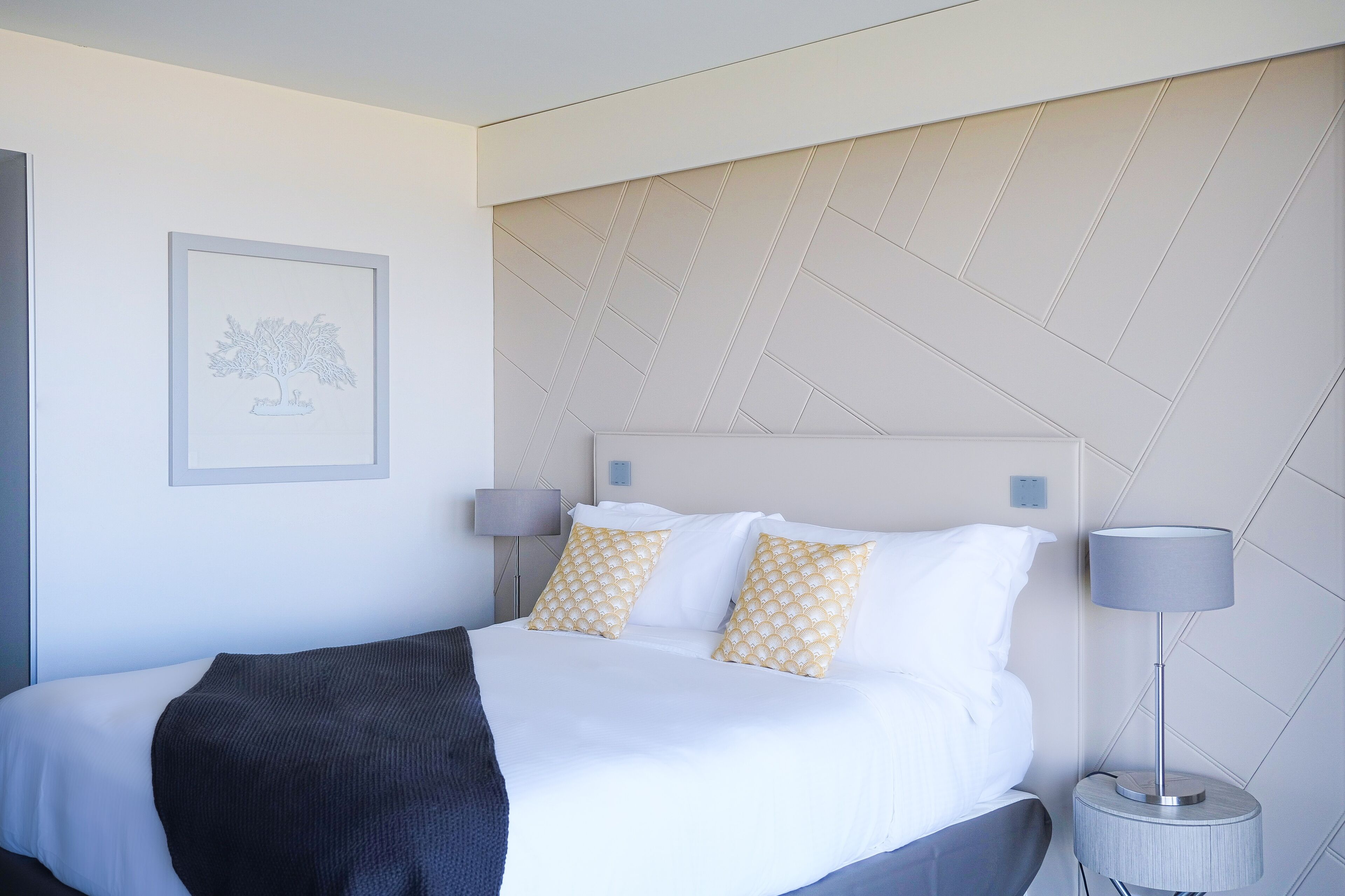A neatly made bed with white and patterned pillows, a dark throw blanket, and a framed picture on the wall.