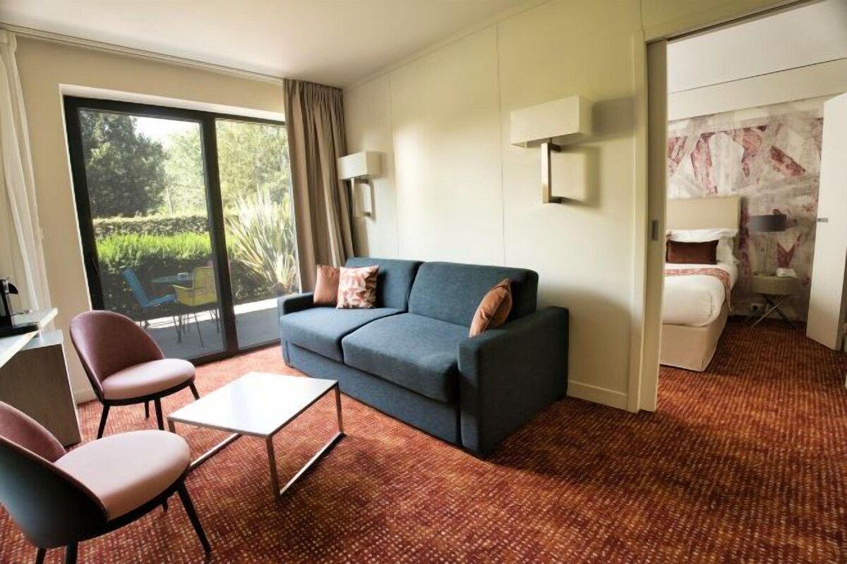 Ein Hotelzimmer mit Sofa, Sesseln, einem kleinen Tisch, einem Bett und einem großen Fenster mit Vorhängen.