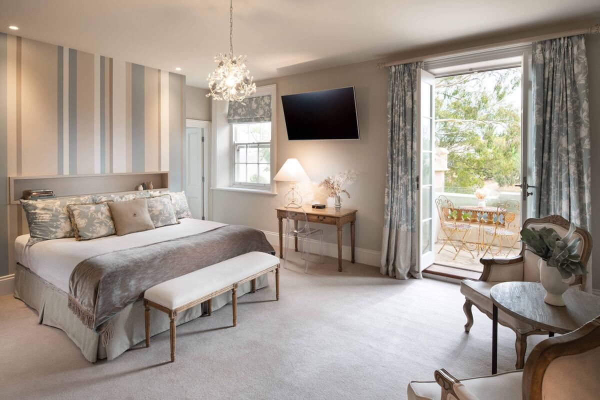 A bedroom with a large bed, a desk, a chair, a TV, and a balcony with a table and chairs.
