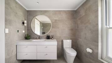 A modern bathroom with a white vanity, a round mirror, a toilet, and a large window.