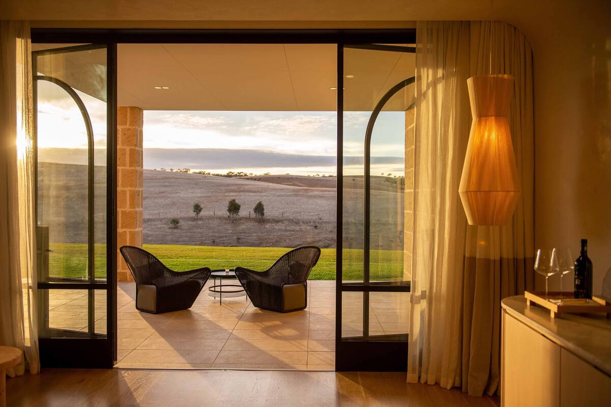 A room with a large glass door leading to a terrace with two chairs and a table, a lamp, and a bottle on a counter.