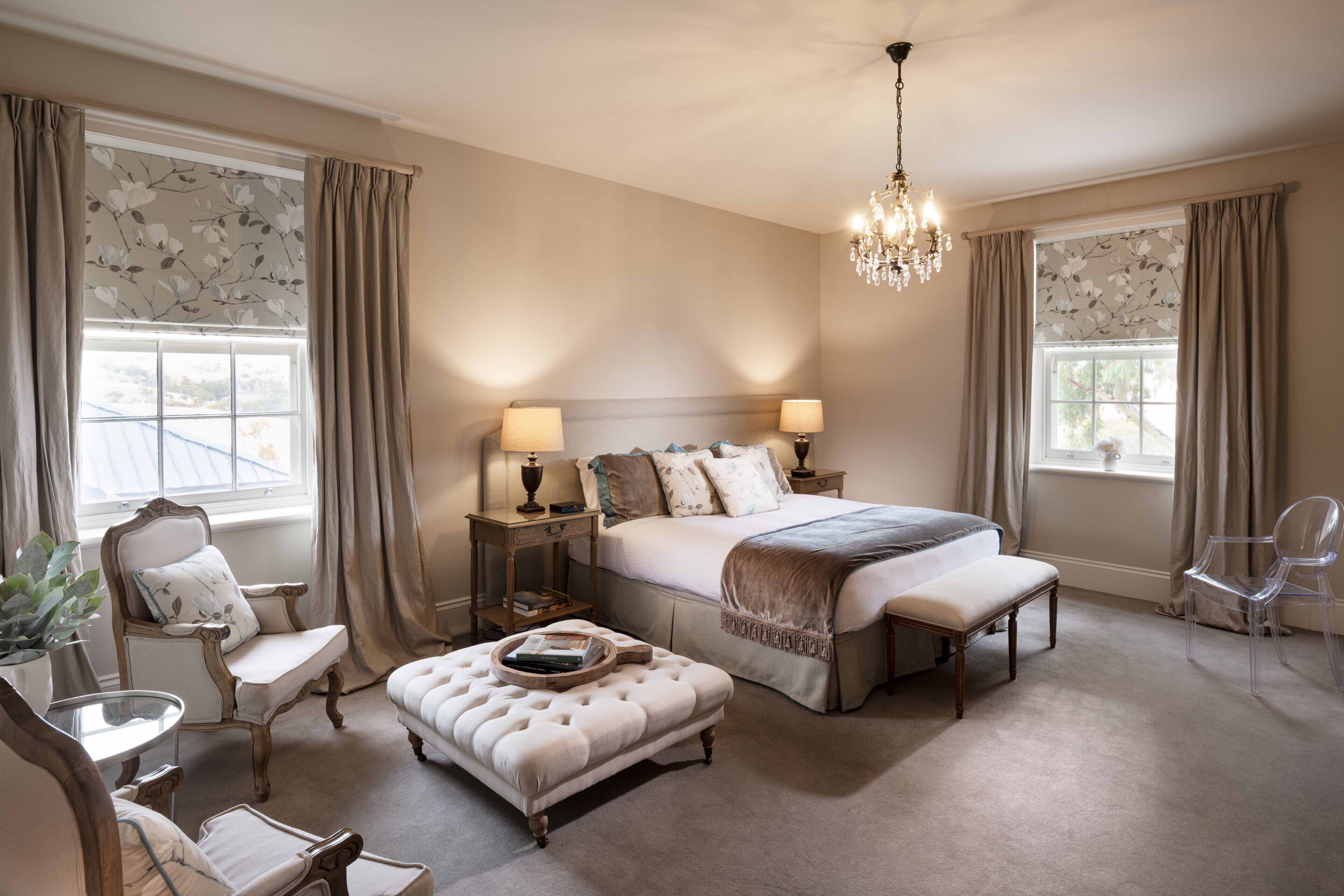 A bedroom with a large bed, two bedside tables with lamps, a chair, an ottoman, and a chandelier.