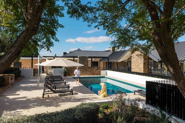Un área de piscina moderna al aire libre con un edificio de piedra, sillones reclinables y una persona caminando.