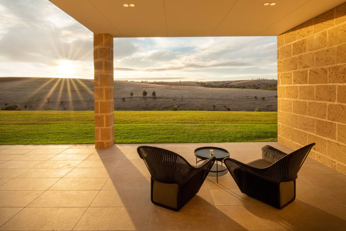 An outdoor patio with two black wicker chairs and a small round table, overlooking a grassy field with distant hills and a clear sky.