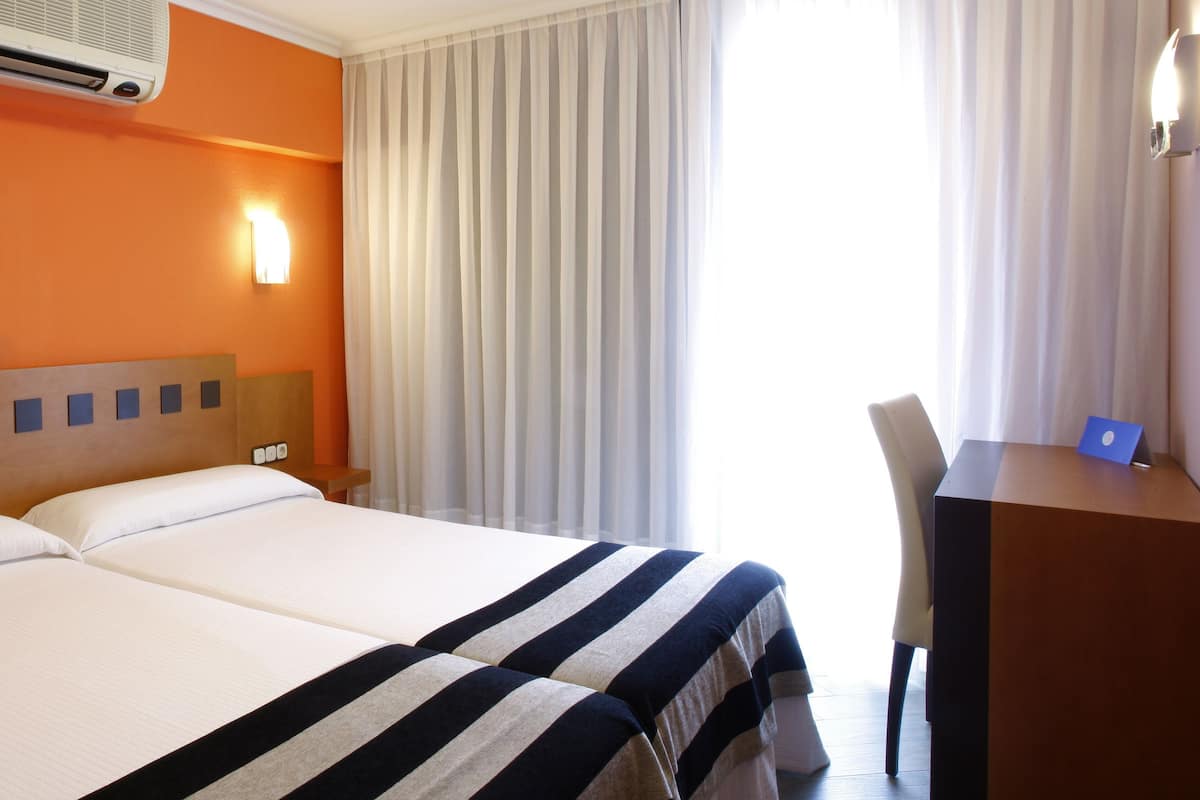 A hotel room with a double bed, a desk with a laptop, an air conditioner, a phone, and a window with curtains.