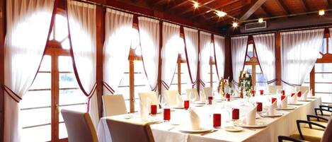 Un comedor con una mesa larga dispuesta para varios invitados, vigas de madera en el techo y ventanas grandes con cortinas.