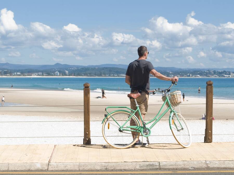 Un uomo in piedi accanto a una bicicletta verde su una spiaggia, con cielo sereno e montagne all'orizzonte.