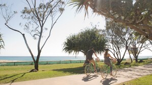 A pathway with two people riding bicycles, a beach in the background, and a palm tree. - La Costa Motel (Bilinga)