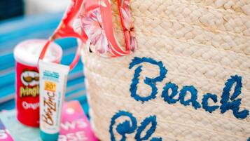 A woven beach tote bag with "Beach Please!" embroidered on it, a book titled "The Art of Travel," and a tube of sunscreen.