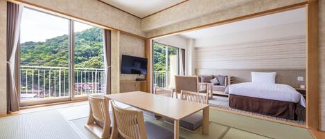 A hotel room with a large window, a bed, a TV, a dining table with chairs, and a sofa.
