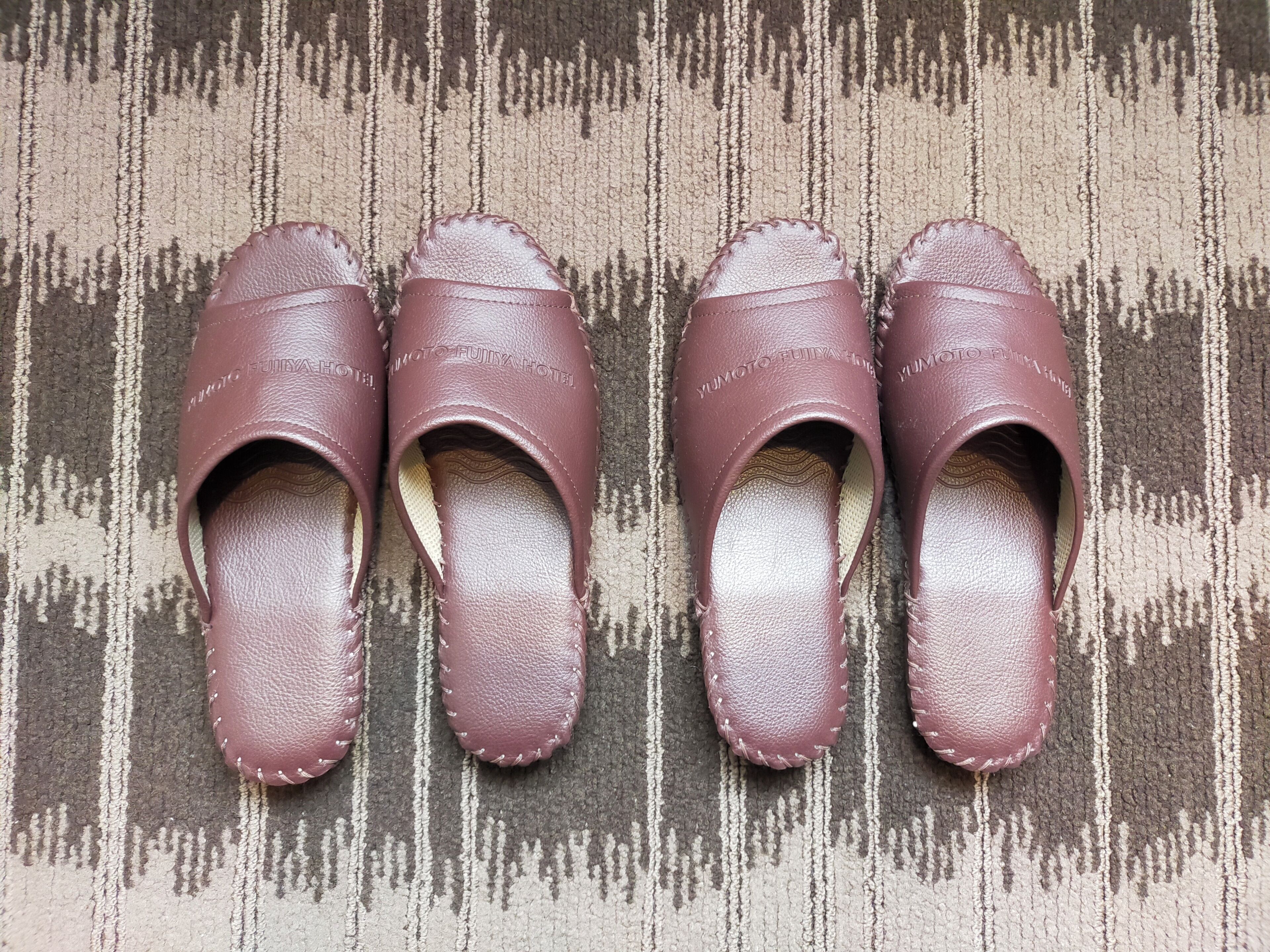 Een paar donkerrode leren slippers met witte stiksels op een kamerbreed tapijt.