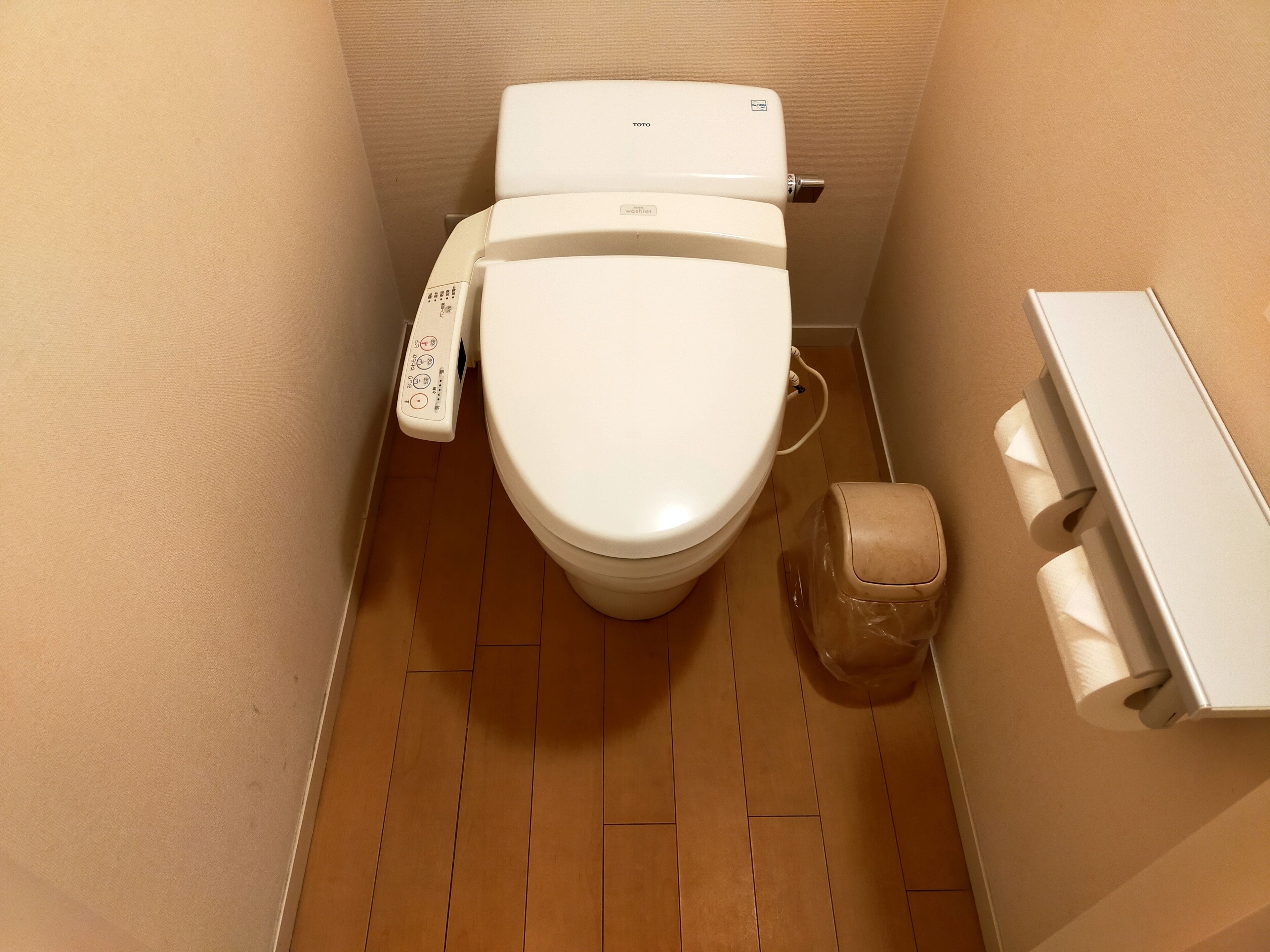 A small, compact bathroom with a white toilet, a toilet paper holder, and a plastic trash bin.