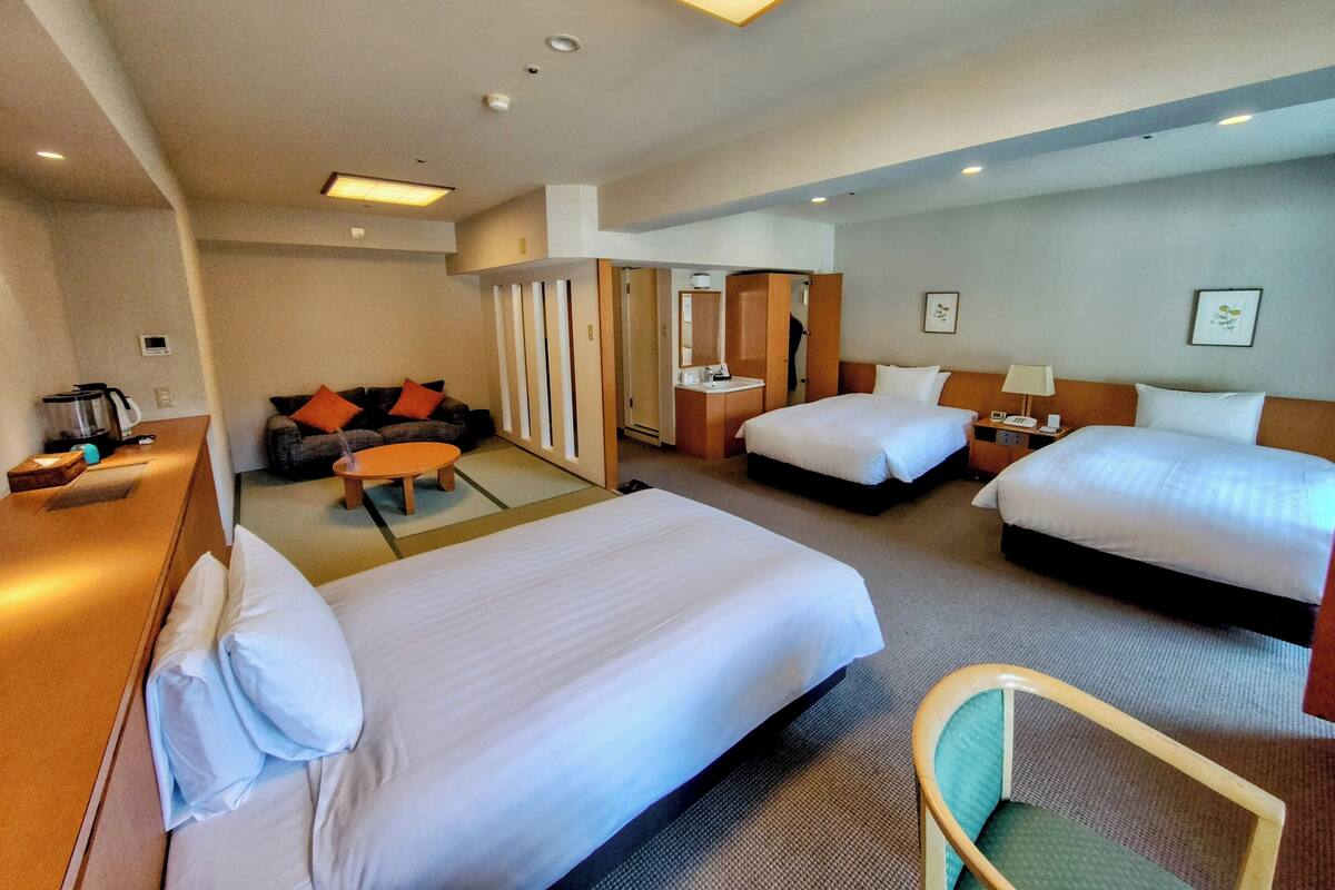 Quarto de hotel com duas camas, um sofá, uma escrivaninha com chaleira e uma cadeira.