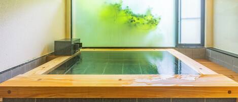 Ein Whirlpool mit Holzkante und gefliestem Boden, ein Fenster mit Blick ins Grüne sowie eine wandmontierte Leuchte.