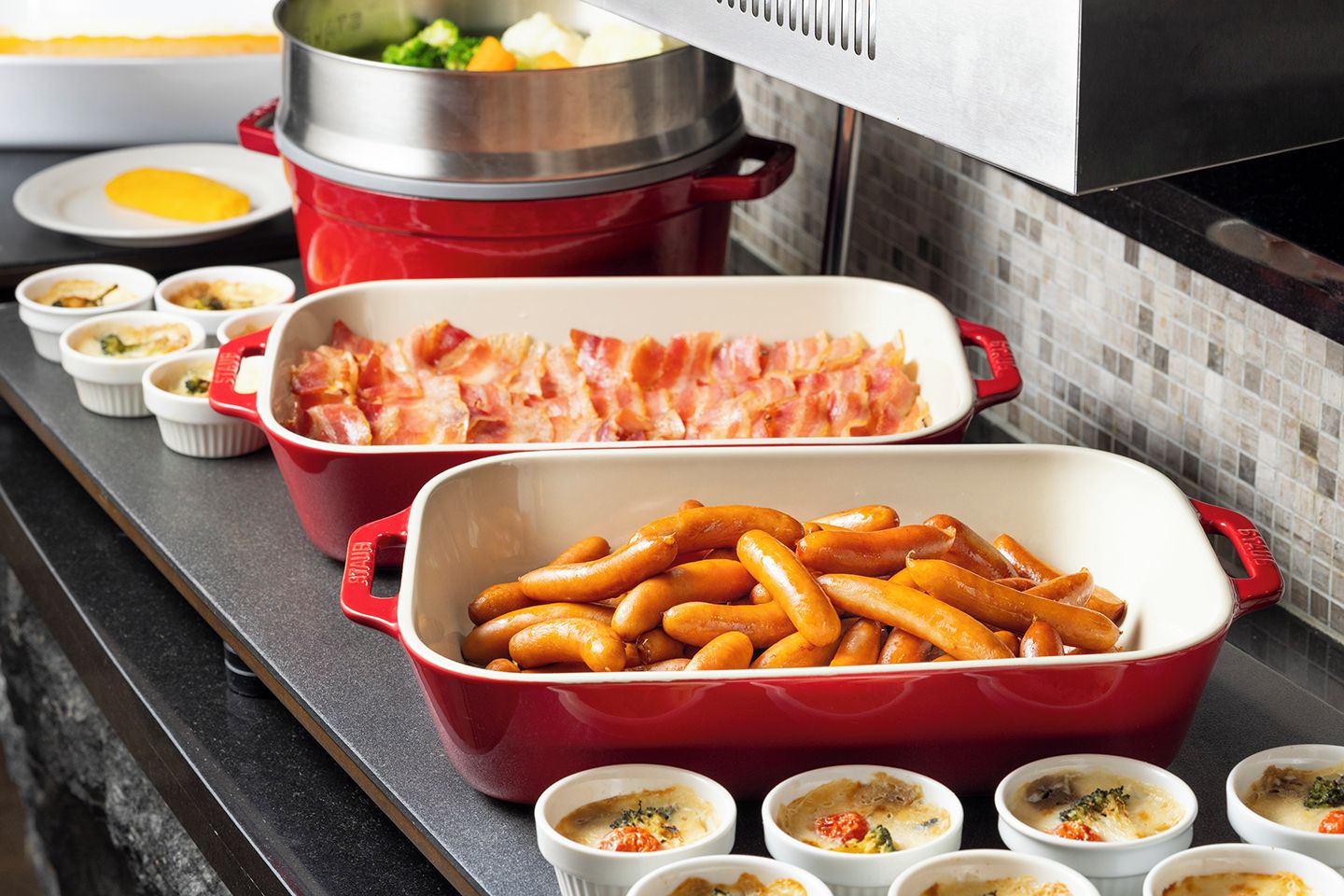 Una encimera de cocina con varios platos, incluyendo una fuente roja con salchichas, una olla con verduras y varios ramequines con distintas salsas.