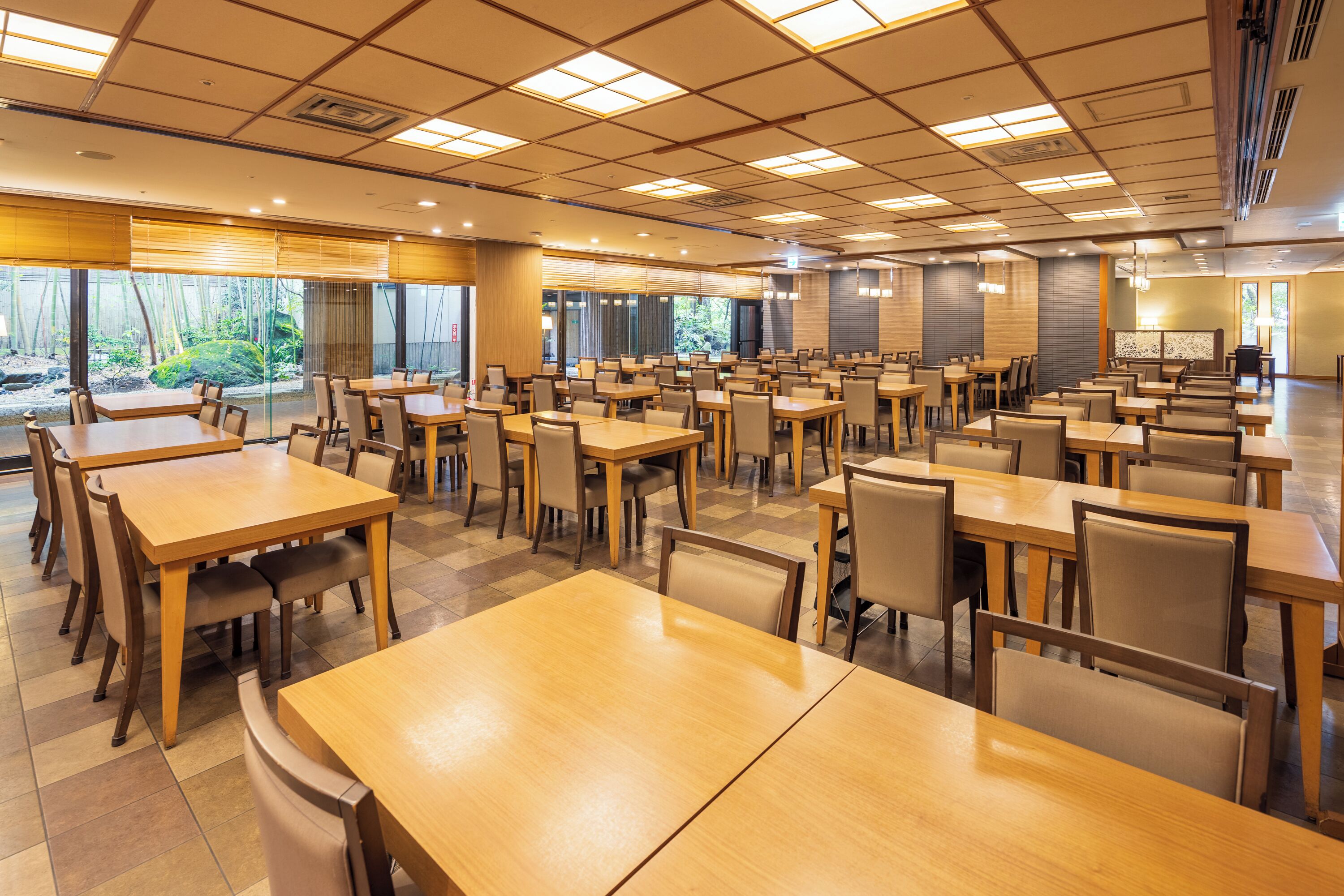 A spacious dining area with multiple tables and chairs, large windows with a view of greenery, and a modern ceiling design.