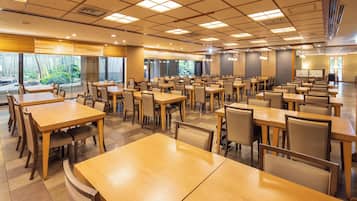 A spacious dining area with multiple tables and chairs, large windows with a view of greenery, and a modern ceiling design.